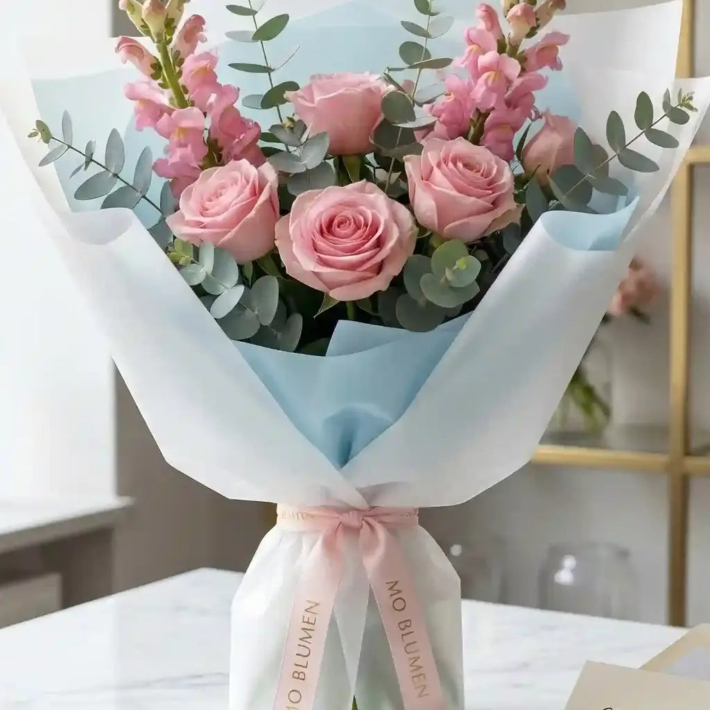 Rosafarbener Rosenstrauß von MO BLUMEN, arrangiert mit rosa Blüten, Eukalyptuszweigen und in hellblau-weißes Papier gewickelt, gebunden mit rosa Band.